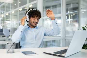 Wall Mural - A cheerful young man with curly hair, wearing headphones, happily listens to music while dancing at his office desk, surrounded by modern technology.