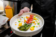 A chef presents an artfully arranged gourmet dish featuring green asparagus, colorful bell peppers, and sauces on a white plate