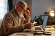 Side view of serious elderly man in eyeglasses checking payment information on receipts while sitting in front of laptop