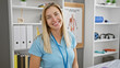 © Krakenimages.com - A cheerful young blonde woman medical professional standing in a clinic room with shelves and anatomy posters.