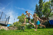 Two kids hold hands, play and jump over a sprinkler in garden
