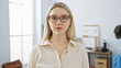 © Krakenimages.com - Portrait of a serious young woman with blonde hair and red glasses standing in a modern office setting