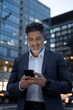 © Stock 4 You - Smiling mature hispanic senior business man using app, holding smartphone cellphone at office building. Indian or latin middle age male entrepreneur businessman working on mobile cell phone. Vertical