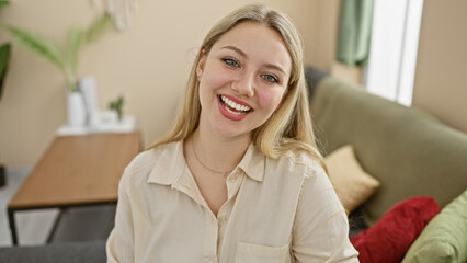 Wall Mural - Smiling blonde woman in a casual beige shirt posing comfortably at her modern furnished living room.