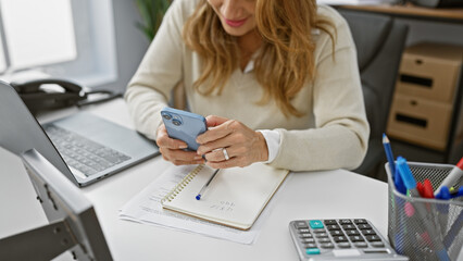 Sticker - A professional hispanic woman uses a smartphone at her modern office desk, surrounded by business essentials.