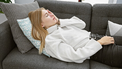 Poster - Blonde woman resting comfortably on a grey couch in a cozy living room, exuding a serene and homely ambiance.