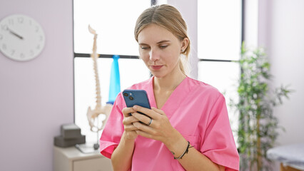 Wall Mural - Blonde woman in pink scrubs using smartphone in a clinic room with medical equipment in the background.