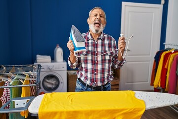 Poster - Cheerful senior man having fun in the laundry room, hilariously sticking out tongue while ironing - truly amusing expression of joy!