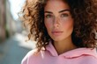 © vejaa - Portrait of young girl with brown curly hair and freckles, no makeup, wearing pink hoodie in city street in sunny day