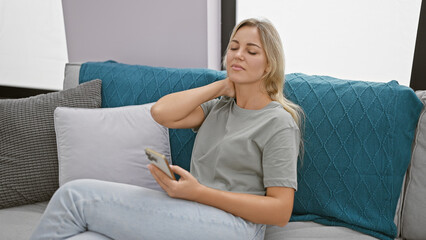 Canvas Print - A young caucasian woman experiencing neck pain while holding a smartphone in a modern living room.