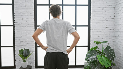 Sticker - Back view of a man standing in a modern room with large windows and a white brick wall, conveying calmness and introspection.