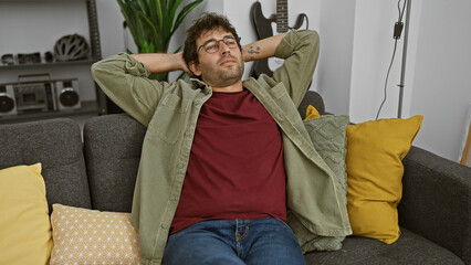 Canvas Print - Relaxed young hispanic man with beard sitting comfortably at home on a gray couch, embodying casual living.