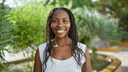 Wall Mural - Smiling young african american woman with curly hair posing for a vibrant portrait in a lush city park.