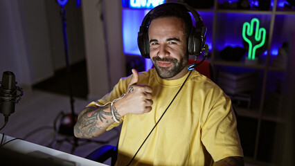 Hispanic man with beard gives thumbs up in a gaming room at night, wearing headphones and a yellow shirt.