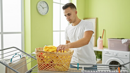 Wall Mural - A young hispanic man folds laundry in a bright green room, beside a washing machine and laundry basket.