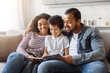 © Prostock-studio - Black family consisting of parents and children are seated together on a couch, engrossed in reading a book. The parents and children are focused on the pages, expressing interest and engagement.