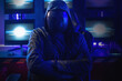 Hacker wearing a hoodie and mask to conceal his identity stands with his arms crossed in front of a monitor, cyber attack, computer security concept