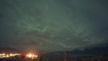 Wall Mural - Northen lights in the cloudy sky above a brightly lit town and port in Northern Norway.