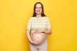 Pretty beautiful friendly young Caucasian pregnant woman dressed in top looking at camera with toothy smile touching her big belly posing against bright yellow wall