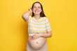Joyful smiling optimistic young Caucasian pregnant woman dressed in top posing against bright yellow wall looking at camera with happy smile gently touching her hair