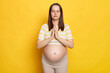 Calm relaxed beautiful young pregnant woman wearing striped top posing isolated over yellow background standing with closed eyes and mudra gesture meditating