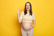 Laughing brown haired pregnant woman with bare belly posing isolated over yellow background showing okay gesture approved sign looking at camera with happy facial expression