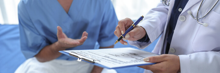 Wall Mural - Professional female doctor with stethoscope around her neck talks with male patient, asks patient to fill out form, patient history, disease symptoms, body aches to plan treatment at hospital bed.