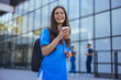 While walking to work in a city hospital, a young female nurse holding coffee to go.  Female medical staff wearing stethoscope relaxing outdoor. Doctor woman drinking coffee.