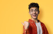 Excited happy black curly haired man in braces, open mouth wear glasses orange shirt advertise show thumb finger area for sales slogan text, isolated yellow background. Dental care ophthalmology ad