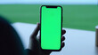 hand holds a smartphone with a green screen in front of a window with a view of a green, grassy field, perfect for apps related to agriculture, landscape design, or environmental monitoring