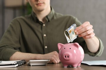 Wall Mural - Financial savings. Man putting dollar banknote into piggy bank at wooden table, closeup