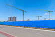 Construction site. Tower cranes behind a blue fence. Residential buildings in the distance. There is a road in the foreground.