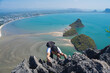 A male hiker ascends a rugged mountain peak with a spectacular view of a serene bay and sandy isthmuses below.