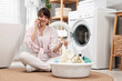 © New Africa - Happy young housewife with laundry talking on smartphone near washing machine at home