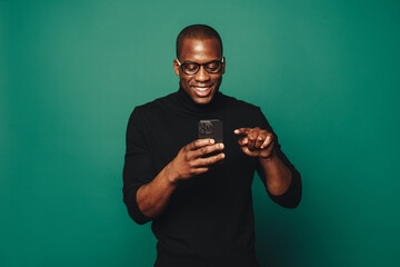Smiling male with stylish glasses holding smartphone and standing against a green background