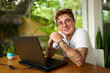 © artiemedvedev - Transgender male person smiles at tropical cafe, laptop on table, earphones nearby. Inclusive work environment, tattoos visible, gender identity embraced, green plants background, youth culture.