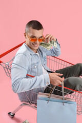 Wall Mural - Young man with money and bags in shopping cart on pink background