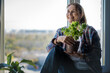 Caucasian attractive middle-aged mature woman standing at the window with a green plant in a pot. Caring for indoor plants