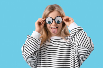 Wall Mural - Little girl in funny eyeglasses showing tongue on blue background. April Fools' Day celebration