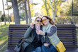 Two happy mature women friends mother and adult daughter taking a selfie on a smartphone while sitting on a bench in the park