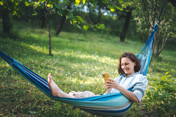 Wall Mural - Cheerful young Caucasian woman in a hammock in a summer garden using a smartphone