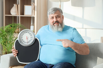 Canvas Print - Overweight happy mature man pointing at scales on sofa at home. Weight loss concept