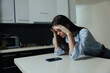© SHOTPRIME STUDIO - Stressed woman sitting on kitchen counter with phone in front of her, feeling overwhelmed and exasperated as she holds her head in hands