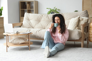 Poster - Young African-American woman with cup of coffee resting on floor at home