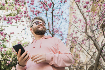 Spring day. Bearded man in pink shirt talking by phone. Spring pink sakura blossom. Handsome young man with smartphone. Fashionable man in trendy glasses. Bearded stylish man. Copy space