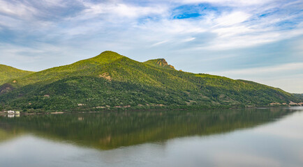  serene landscape featuring a mountain range with lush greenery, reflected in the calm waters of a lake. The sky is partly cloudy, and the overall scene exudes tranquility and natural beauty.