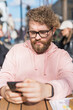 Portrait of cheerful millennial blogger with modern cellphone technology enjoying freelance lifestyle, happy hipster guy in optical eyewear using mobile phone in street cafe