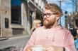 Young bearded man having breakfast at table of street cafe on spring day, drinking warm cappuccino and enjoy morning.