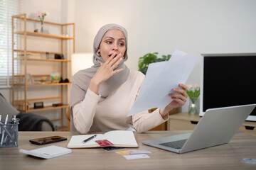 Wall Mural - Young worried and shocked Muslim woman in hijab holding documents and calculator. Financial problems with accounts, loans, family budget at the home