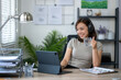 © Wasana - A woman is sitting at a desk with a laptop and a glass of water. She is wearing headphones and she is enjoying her work